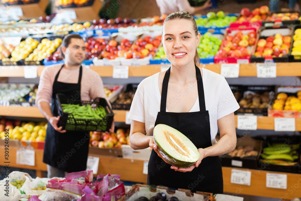 Young female seller with half of melon and man on background having box of green beans on the supermarket