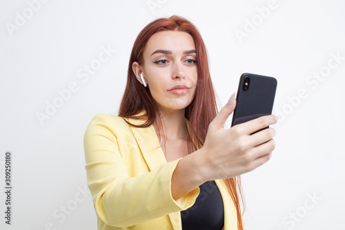 A red-haired girl in a yellow suit listens to music in headphones and takes a selfie on a white background