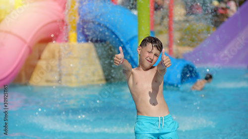 Active Caucasian boy spending his summer holidays in water park, he splashing in pool against colorful plastic slide and showing like gestures. © Artem