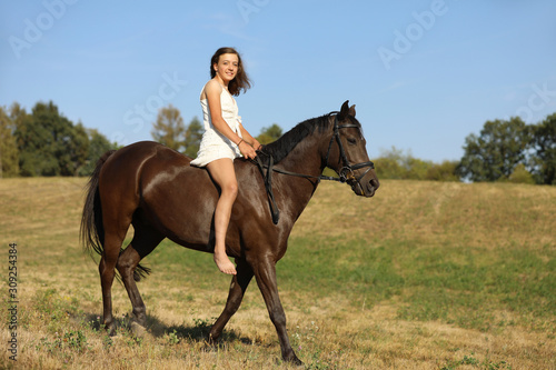 Happy teenage pretty girl on pony without saddle running on meadow in hot summer afternoon