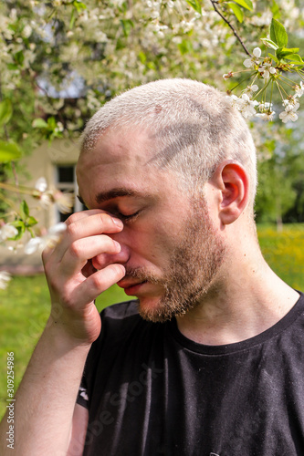 08.05.2019, Moscow, Russia. A man rubbing his nose suffering from the seasonal pollen allergy attack standing next to the flowering tree in the spring park.