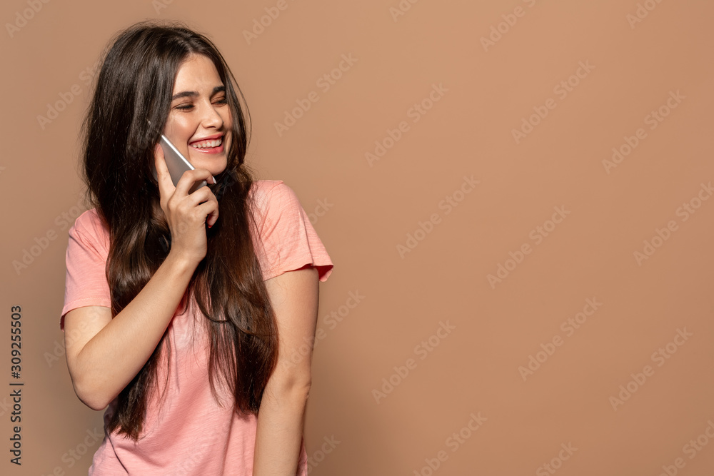 Fototapeta premium Freestyle. Young girl in t-shirt standing studio isolated on brown talking on smartphone laughing joyful copy space