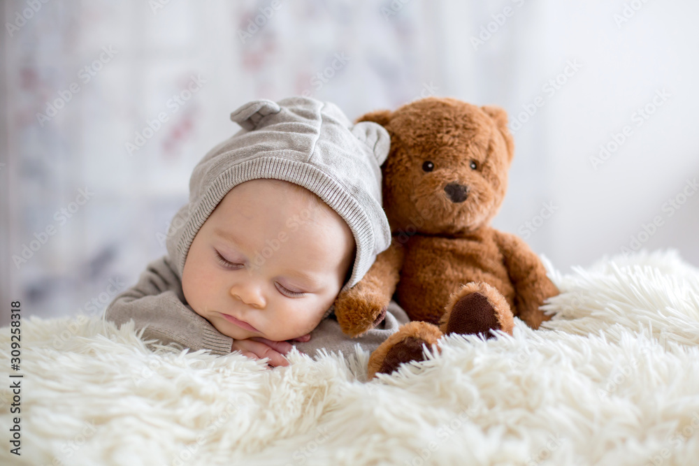 Sweet baby boy in bear overall, sleeping in bed with teddy bear - obrazy, fototapety, plakaty 