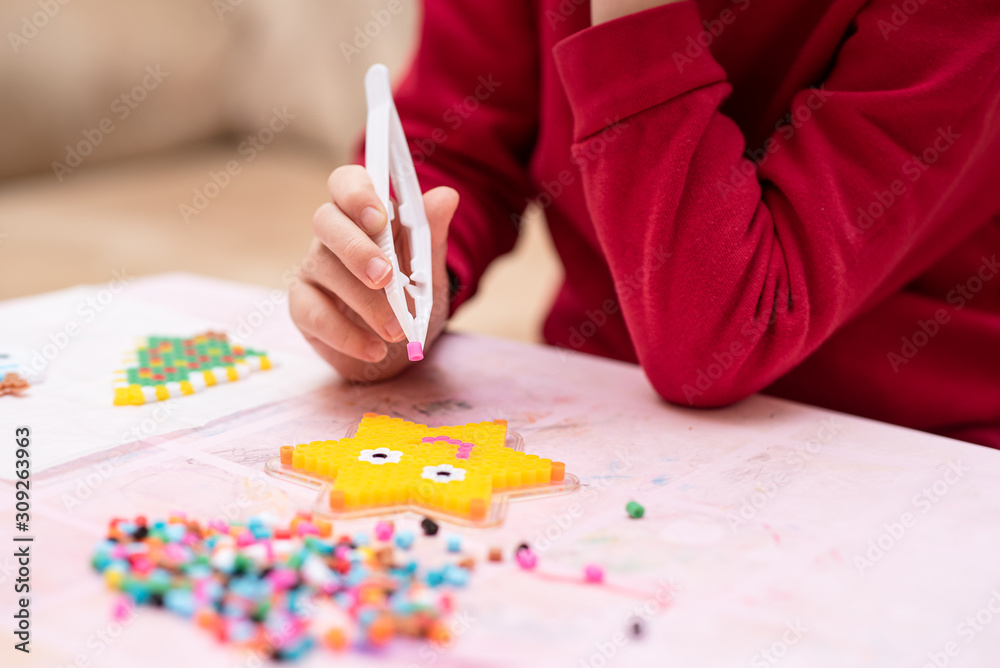 Little girl holds a tweezers with bead, creates art toy from fusible  colorful beads, last touches. Also known as perler beads. Toy that develops  the imagination of child. Process close up Stock