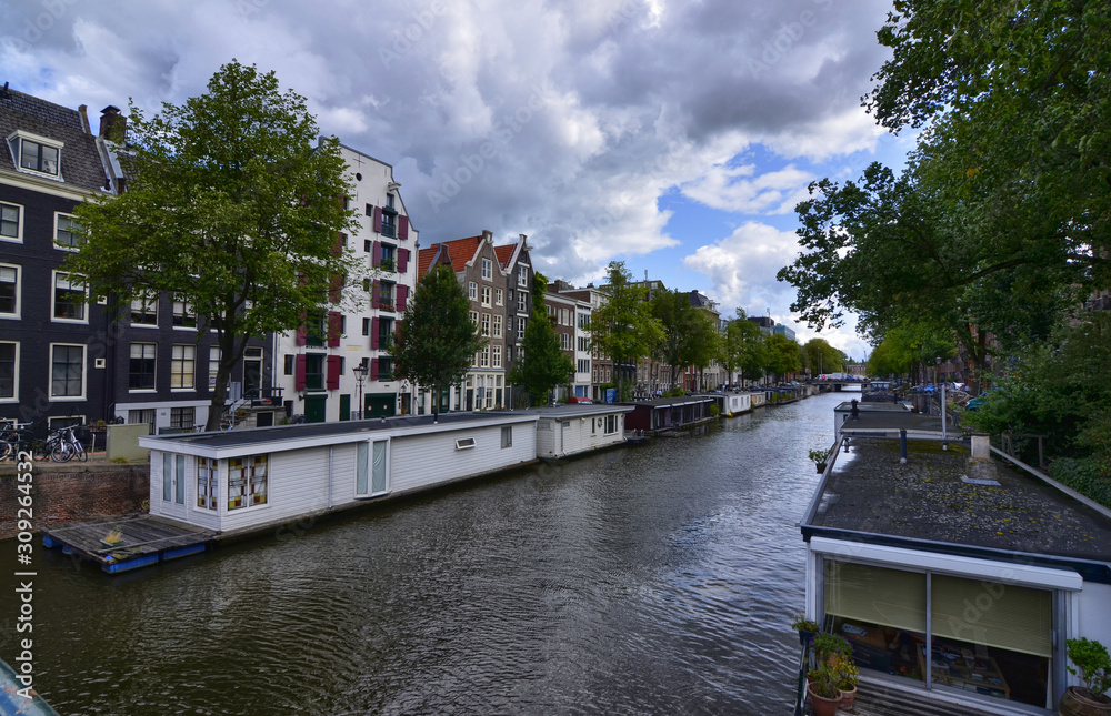 Amsterdam,Holland,August 2019. In the historic center a view that is a symbol of the city: a white houseboat moored on the edge of the canal. Behind it the tree-lined avenue and the little houses