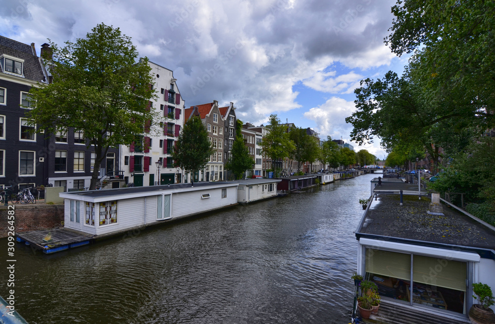 Amsterdam,Holland,August 2019. In the historic center a view that is a symbol of the city: a white houseboat moored on the edge of the canal. Behind it the tree-lined avenue and the little houses