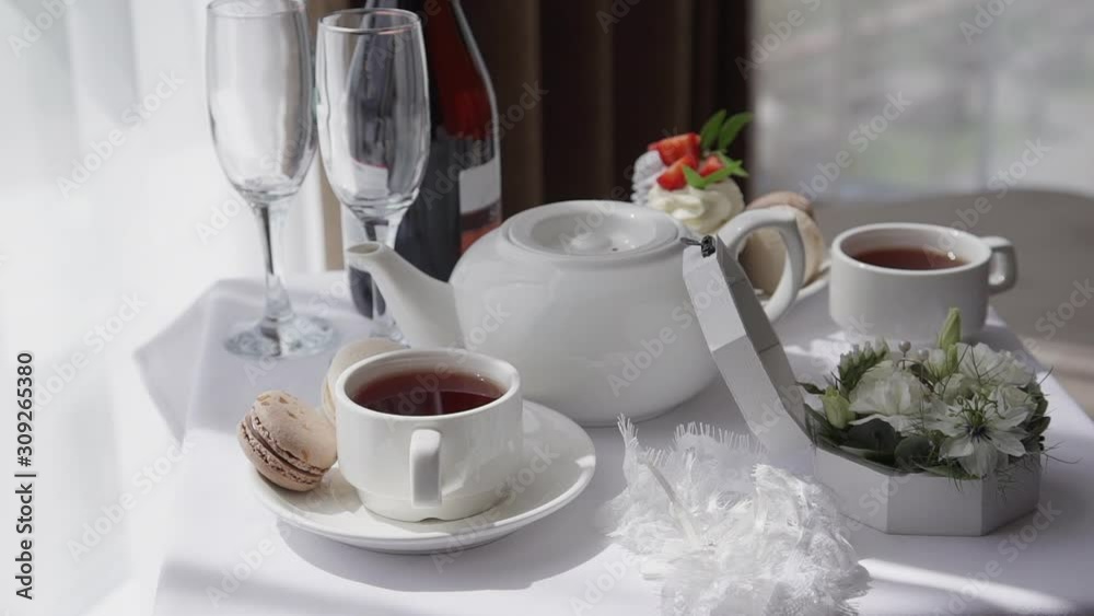Festive table with food and drink for romantic date in luxury hotel interior. Tea, wine bottle, flowers and light macaroon are on white desk in light room by window. Concept: luxury, arrangement Festive table with food and drink for romantic date in luxury hotel interior. Tea, wine bottle, flowers and light macaroon are on white desk in light room by window. Concept: luxury, arrangement