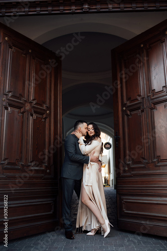Couple standing at the entrance to a large hall near a large open wooden door. Happy girl in dress and guy posing in suit. Beautiful plus size model in dress outdoors, xxl woman. Happy to be together.