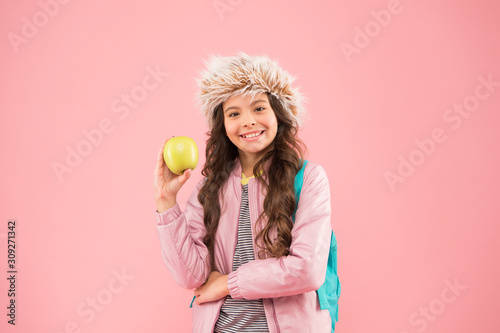end of winter semester. holiday and vacation. kid pink backdrop. teen daily life. education and happy people schoolgirl with books after lesson. school break. small happy girl earflap hat eat apple