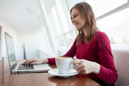 young girl freelancer sitting in a cafe with a laptop and drinking coffee, woman use a computer near the window