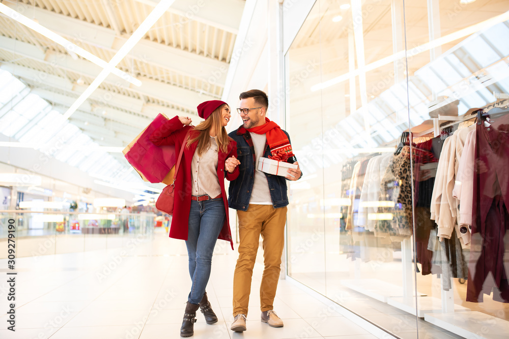 Happy caucasian couple enjoying Christmas shopping and looking each other while standing by the shop window in a mall. Girlfriend is carrying shopping bags and boyfriend is holding Christmas gift.
