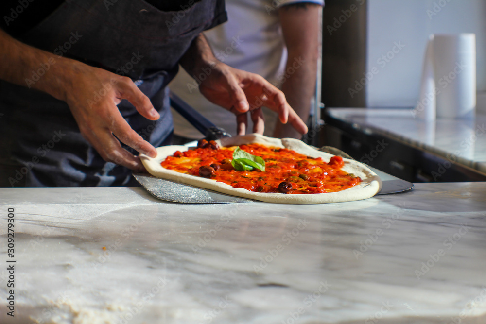 pizza maker is preparing a Gourmet pizza with San Marzano tomato sauce, black olives, capers, fresh basil, EVO oil and a sprinkling of pecorino in an Italian pizzeria