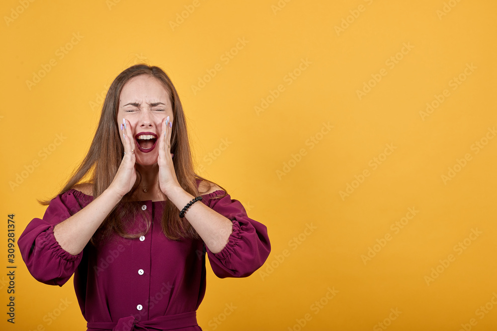 Young woman with burgundy bluse over isolated orange background shouting with mouth wide open. Girl holding hands near mouth Young woman with burgundy bluse over isolated orange background shouting with mouth wide open. Girl holding hands near mouth