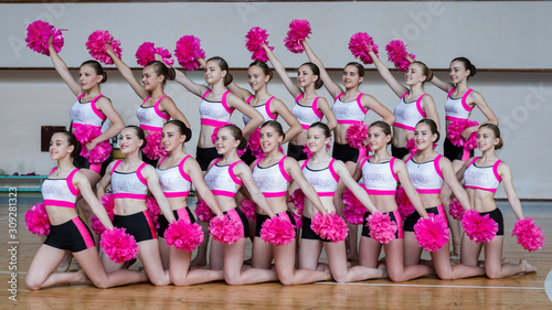 attractive young cheerleaders working out in sports club, group of cheerleaders with pom-poms in their hands, hands raised up, girls in black and pink suit with pompons on the background of the gym