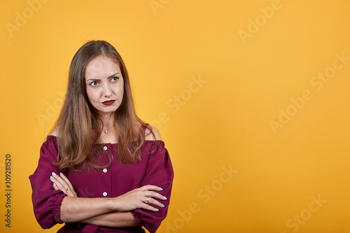 Ginger girl in burgundy bluse with bow over isolated orange background thoughtfully looks right folding her arms