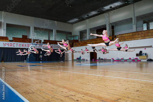 active pretty girls in action, group of cheerleaders jumping in the air demonstrate perfect stretching, hands raised up, girls in black and pink suit on the background of the gym, horizontal twine