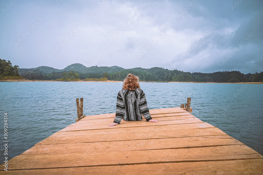 Young woman sitting on a pier watching a lagoon with mountains in the background and cloudy sky Young woman sitting on a pier watching a lagoon with mountains in the background and cloudy sky