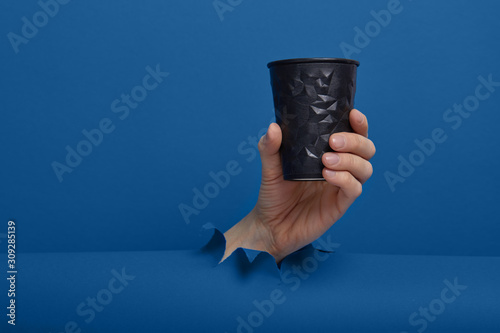 Male hand taking a cup of coffee through torn classic blue paper background. Get energy boost, hot drink in take-away package. photo