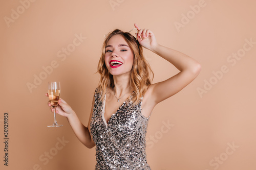 Close-up portrait of pretty white girl with curly hairstyle posing at festive with excitement. Joyful young woman with goblet standing with hand up and laughing on beige background.