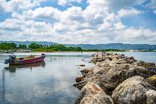 Empty old wooden fishing motor boat docked near shore. Scenic ocean water view setting from the north coast of Montego Bay, Jamaica, with large sea boulders. Trees and mountains in the background.  photo