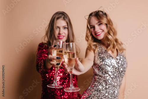 Indoor blur portrait of spectacular young ladies with wineglasses in focus. Pretty girl with blonde curly hairstyle celebrating birthday with her best friend in red dress.