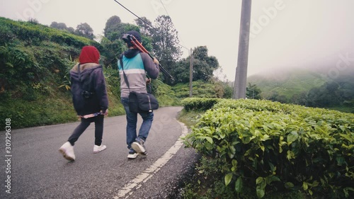 Young couple, enjoying view at tea farm, Genting Highlands, adventure journey, photographer carrying tripod, walking together with partner at rural meadow nature landscape at morning.