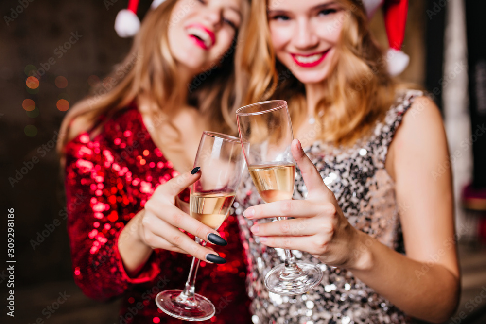 Indoor close-up photo of two winsome girls in new year hats celebrating holidays together. Portrait of funny ladies clinking glasses of champagne in christmas day.