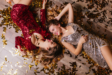 Overhead portrait of spectacular european girls lying on confetti during party. Charming ladies in glamorous clothes resting after dance at new year event.