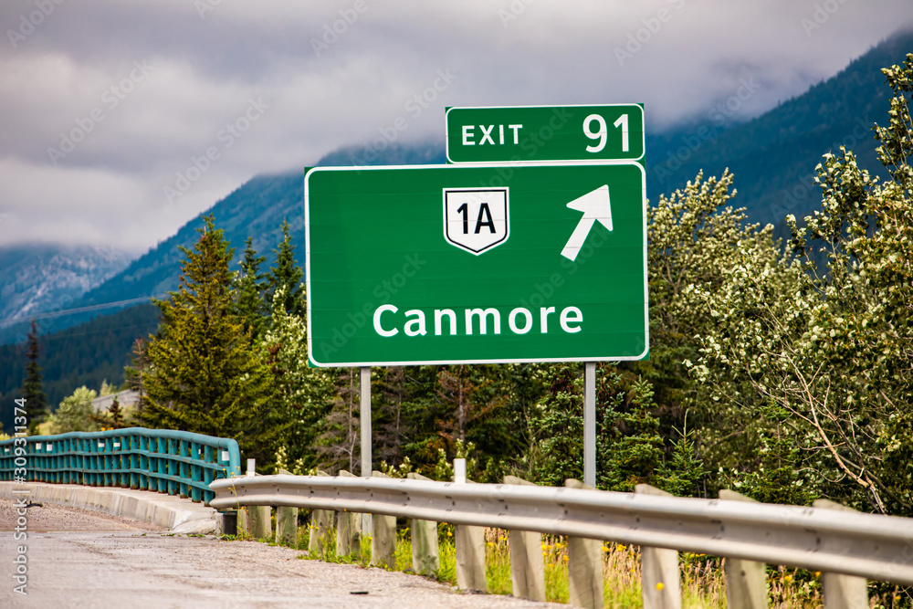 Fototapeta premium Information road green sign, the next exit after 91 km, Canmore, 1A road symbol, Canadian rural roadside forests in the background, Alberta, Canada