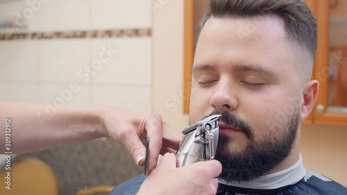 Stylist using clipper and black comb to cut males beard, close shot. Beige interior of hairdressing saloon. Man in barbershop. Selective soft focus. Blurred background