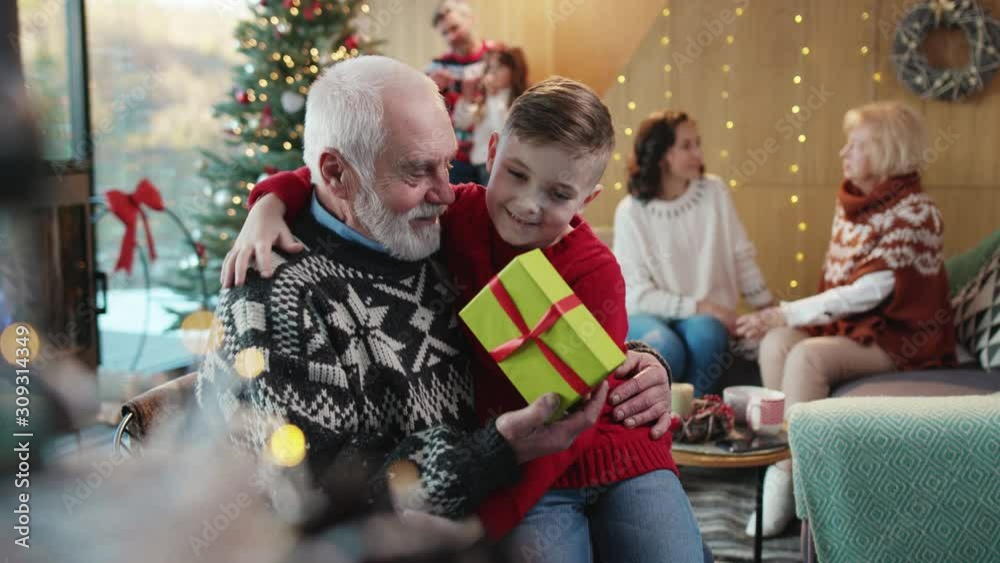 Happy family celebrating magic Christmas at home. Cheerful cute boy giving a surprise gift for grandpa, hugging and sitting together in beautiful decorated holiday room. Happy family celebrating magic Christmas at home. Cheerful cute boy giving a surprise gift for grandpa, hugging and sitting together in beautiful decorated holiday room.