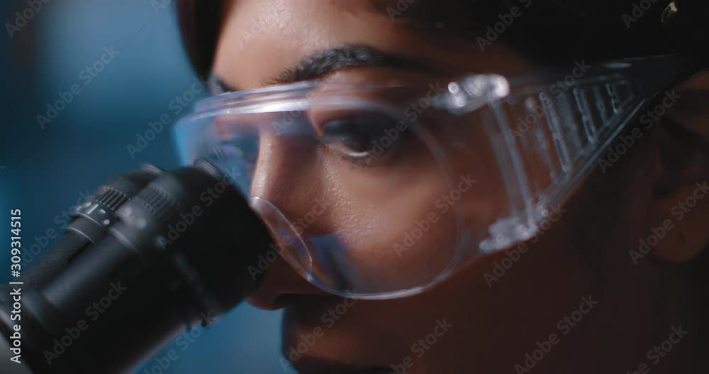 Close up of a female research scientist eyes, looking at samples under microscope, wearing safety glasses.Slow motion, slider, shot with BMPCC 4K.Biochemistry, pharmaceutical medicine,science