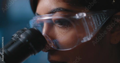 Close up of a female research scientist eyes, looking at samples under microscope, wearing safety glasses.Slow motion, slider, shot with BMPCC 4K.Biochemistry, pharmaceutical medicine,science