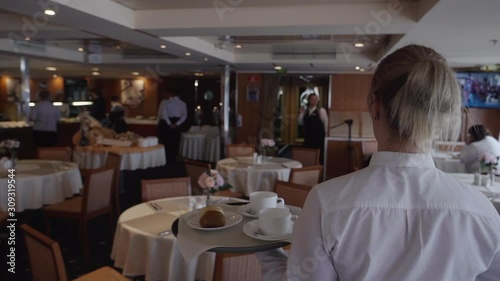 Female waiter carries food and cups on tray while work in restaurant. Back view of woman worker wearing white uniform is walking and working in banquet hall. Decorated tables and staff in interior