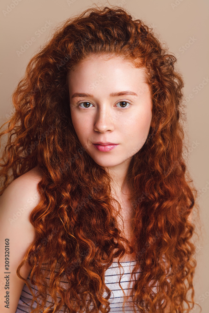 Beautiful teenage girl with red curly long hair and brown eyes without makeup. Portrait shot in studio on a beige background. Retouched skin. Beautiful teenage girl with red curly long hair and brown eyes without makeup. Portrait shot in studio on a beige background. Retouched skin.