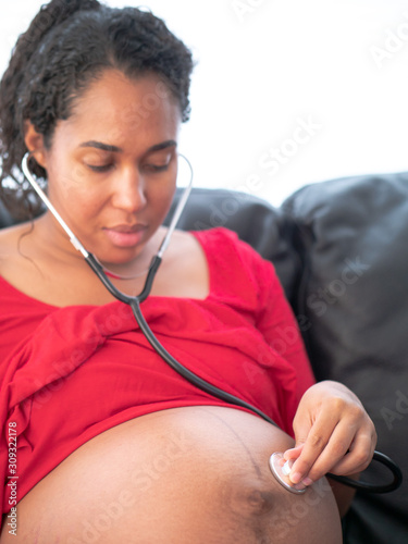 A beautiful young African American mixed race woman with dark curly hair in a pony tail and red dress listens to her unborn baby's heart beat using a stethoscope on her bare skin belly.