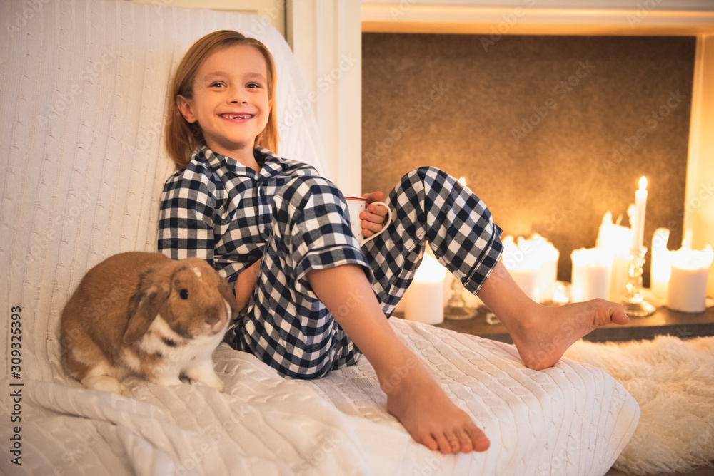Smiling cheerful little blond boy sitting on a couch in front of the fireplace, with little rabbit Smiling cheerful little blond boy sitting on a couch in front of the fireplace, with little rabbit