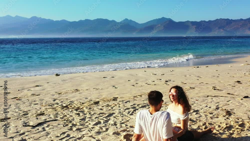 Young couple sitting on white sand of exotic beach, talking about travel and tour around tropical islands in Indonesia during their vacation Young couple sitting on white sand of exotic beach, talking about travel and tour around tropical islands in Indonesia during their vacation
