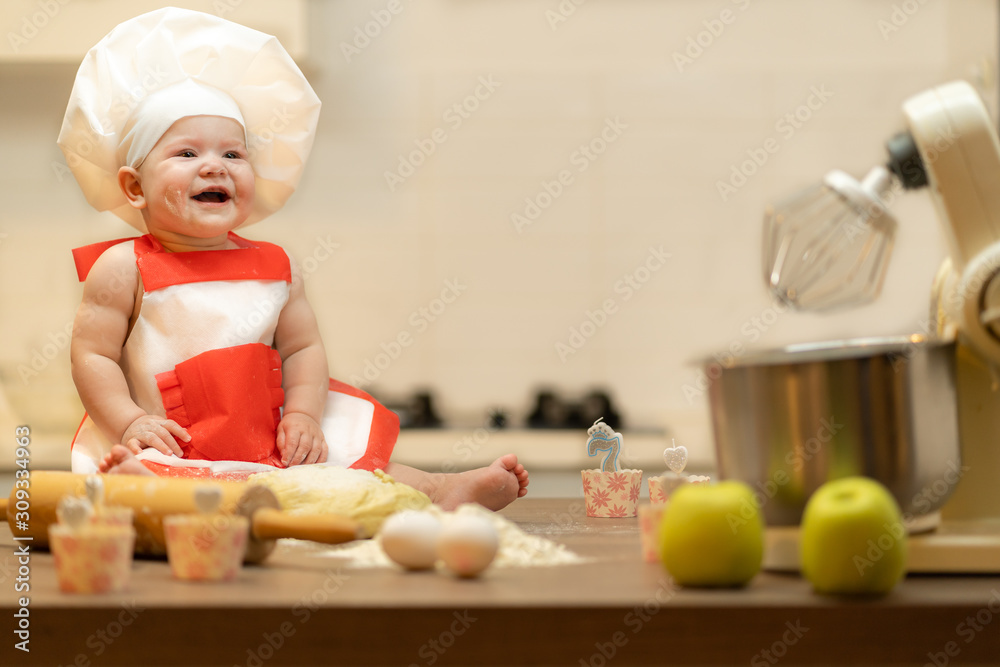 A Child, Baby, Girl, sits On The Kitchen Table, In A Cook's Cap A Child, Baby, Girl, sits On The Kitchen Table, In A Cook's Cap