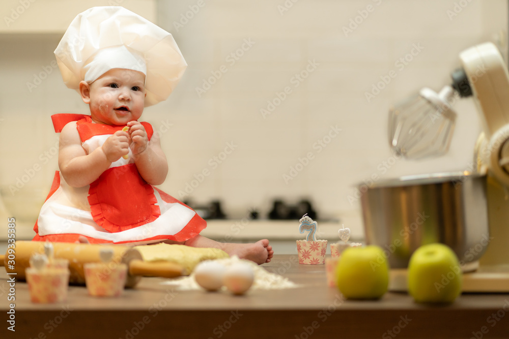 A Child, Baby, Girl, sits On The Kitchen Table, In A Cook's Cap A Child, Baby, Girl, sits On The Kitchen Table, In A Cook's Cap