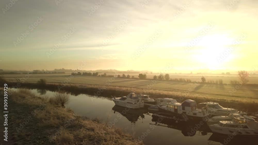 Drone flies towards sunrise over river with boats moored up and frosty fields. Reflections on surface of the water and big shadows cast into fields by trees. Drone flies right and camera pans left. Drone flies towards sunrise over river with boats moored up and frosty fields. Reflections on surface of the water and big shadows cast into fields by trees. Drone flies right and camera pans left.