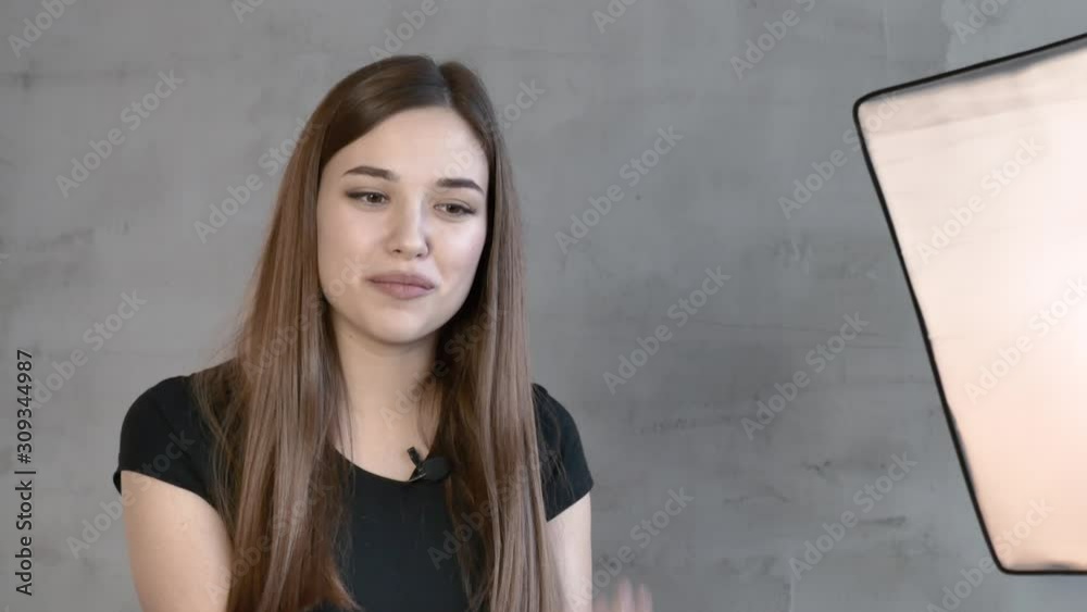 Close up. Caucasian attractive Girl in a black t shirt with long straight hair brown haired against a gray wall and a lighting fixture speaks to the camera gestures Close up. Caucasian attractive Girl in a black t shirt with long straight hair brown haired against a gray wall and a lighting fixture speaks to the camera gestures