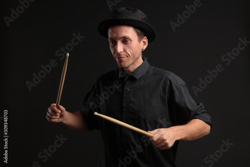 Stylish man drummer in a black shirt and hat playing drums with sticks over dark background.