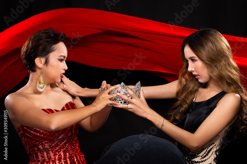 Two Miss Beauty Pageant Queen Contestants fight, catfight, for a jewel Diamond Crown. They wear Sequin Long Gown and struggle for the prize in the background of red cloth flying in the Air photo