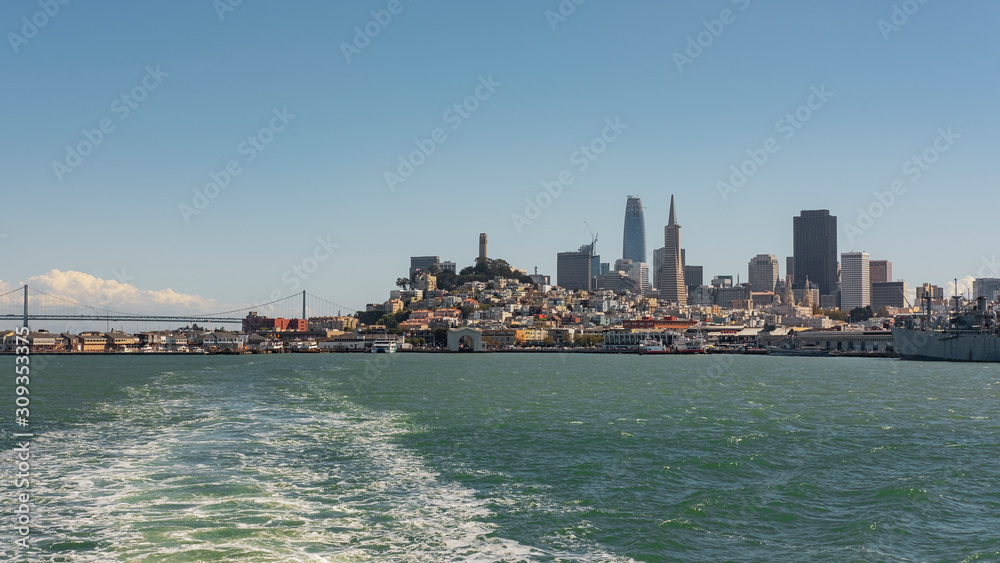 Stunning vistas of San Francisco observed from the stern of a ferryboat departing the pier with frothy trail, towards the downtown financial district, the bay and Oakland Bay Bridge, in California, US