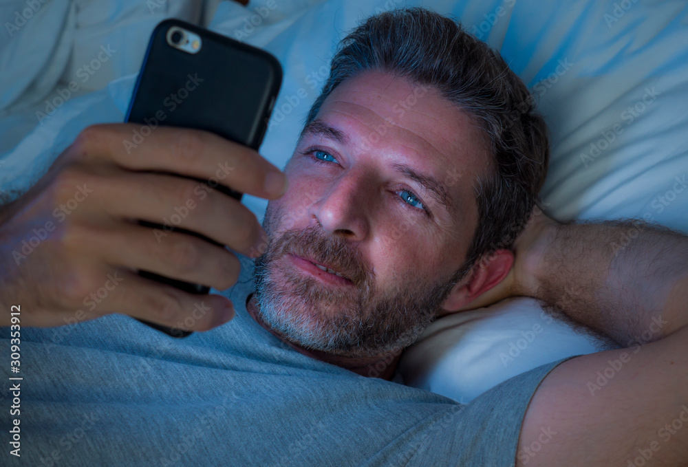 Obraz premium happy man with blue eyes lying on bed late at night in dark light networking on mobile phone or online dating smiling relaxed enjoying internet in social media addiction
