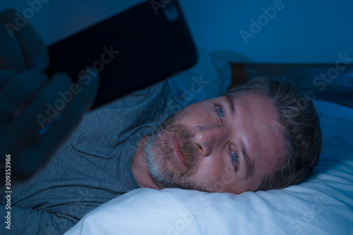 happy man with blue eyes lying on bed late at night in dark light networking on mobile phone or online dating smiling relaxed enjoying internet in social media addiction