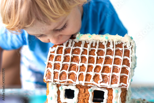 Сute little boy eats gingerbread house and having fun. Kid enjoy Christmas traditions. Child broke a gingerbread house. Baking and cooking with children for Xmas at home. Cristmas concept. photo