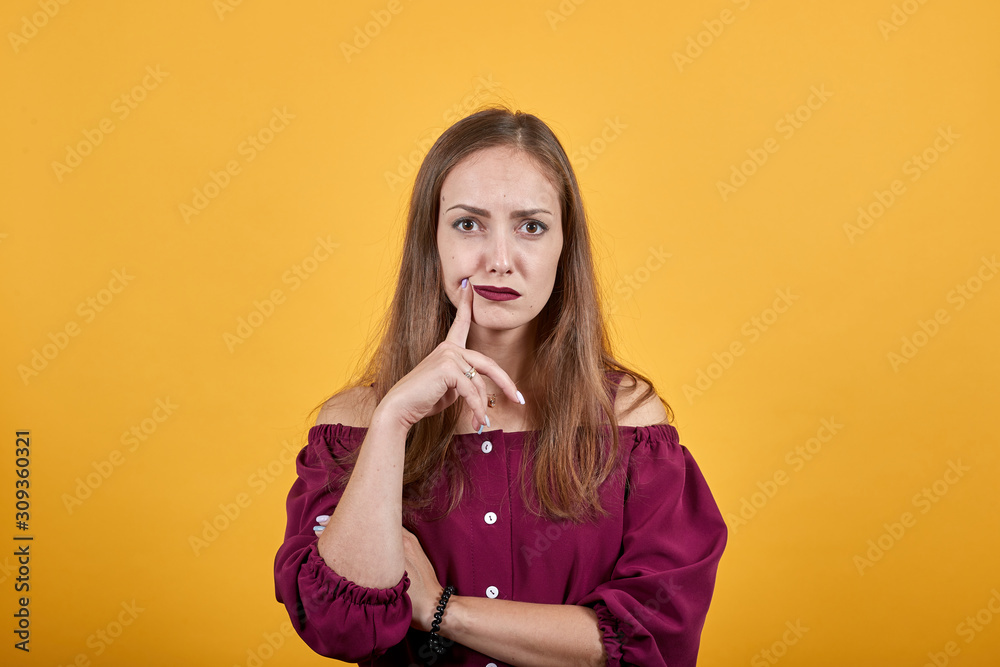 Ginger girl in burgundy bluse with bow over isolated orange background thoughtfully looks straight hold her finger on cheek Ginger girl in burgundy bluse with bow over isolated orange background thoughtfully looks straight hold her finger on cheek