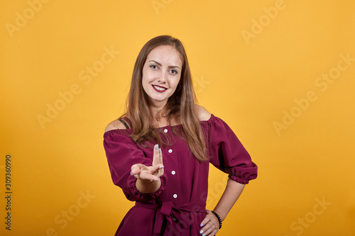 Young girl standing and inviting to come with hand. She is dressed up in burgundy bluse with bow, behind her there is orange wall. She looks open-hearted